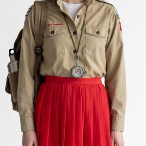 May include: A tan long-sleeved button-down shirt with a Scouts America patch, a red pleated midi skirt, and a tan backpack with a silver water bottle. A round compass hangs from a cord around the neck over the shirt, which is worn over a white t-shirt.