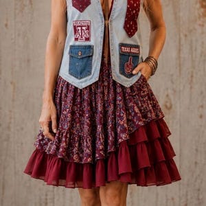 Peut inclure: Un gilet sans manches en jean bleu clair avec des poches à motifs rouge foncé et des écussons "A&M" et "TEXAS AGGIES" porté sur une robe bordeaux arrivant aux genoux avec un imprimé floral et un ourlet à volants, avec des bottes de cow-boy en cuir marron.