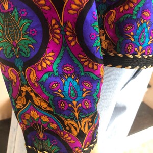 May include: Close-up of a sleeve with a vibrant, ornate pattern. The design features a mix of purple, teal, gold, and green floral and geometric shapes on a black background. The cuff is trimmed with black fabric and gold stitching.