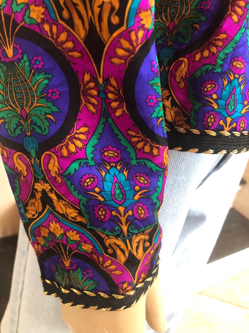 May include: Close-up of a sleeve with a vibrant, ornate pattern. The design features a mix of purple, teal, gold, and green floral and geometric shapes on a black background. The cuff is trimmed with black fabric and gold stitching.