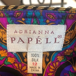 May include: Close-up of a colorful silk garment with a label that reads "ADRIANNA PAPELL". The fabric features a vibrant floral pattern in shades of purple, teal, and gold. The label indicates the garment is 100% silk, size 12, and made in China.