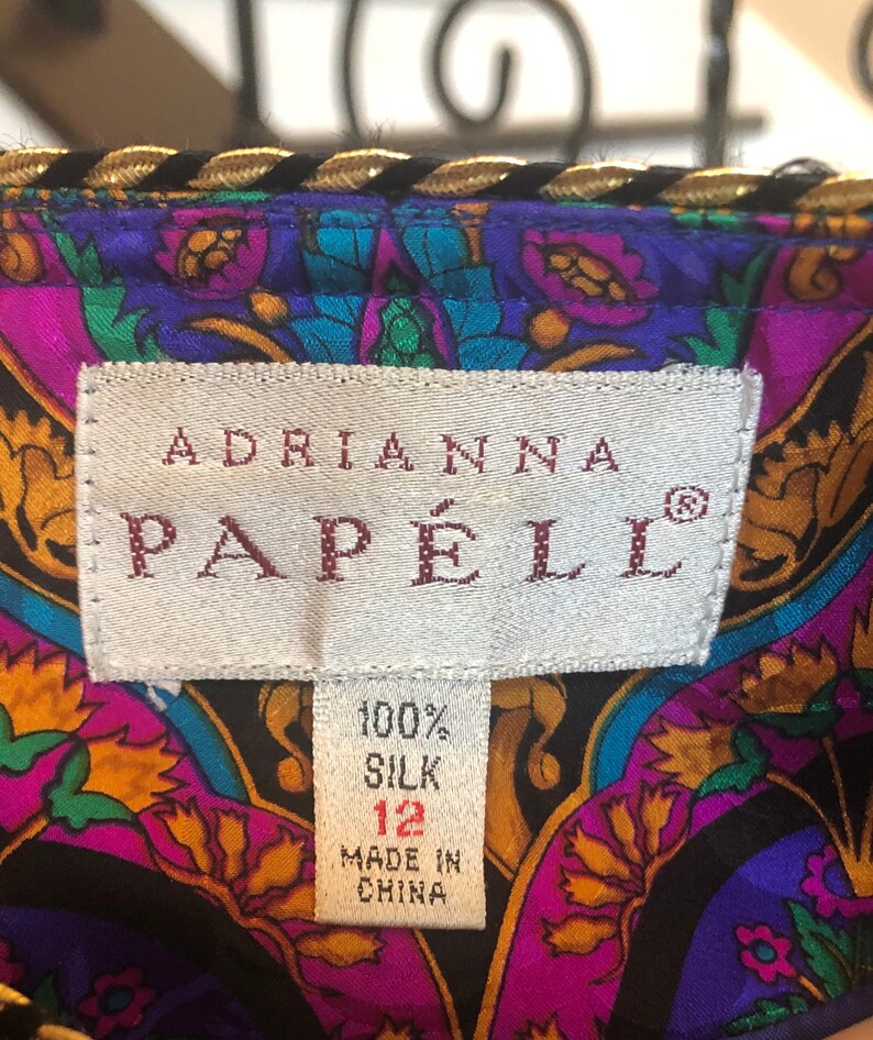 May include: Close-up of a colorful silk garment with a label that reads "ADRIANNA PAPELL". The fabric features a vibrant floral pattern in shades of purple, teal, and gold. The label indicates the garment is 100% silk, size 12, and made in China.