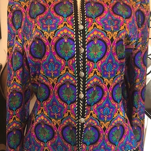 May include: A vibrant, patterned jacket in shades of purple, blue, and gold. The jacket features a black trim with silver buttons and a decorative design. The long-sleeved jacket is displayed on a mannequin, showcasing its intricate details and colorful pattern.