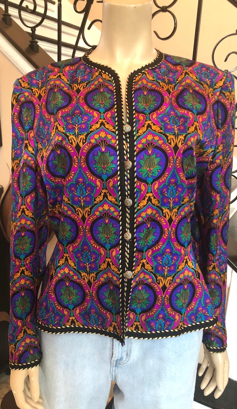 May include: A vibrant, patterned jacket in shades of purple, blue, and gold. The jacket features a black trim with silver buttons and a decorative design. The long-sleeved jacket is displayed on a mannequin, showcasing its intricate details and colorful pattern.