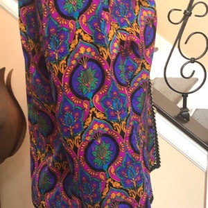 May include: A patterned jacket with a vibrant design in purple, blue, gold, and green. The jacket has a black trim and is worn over light blue jeans. The jacket's pattern features a repeating floral motif.
