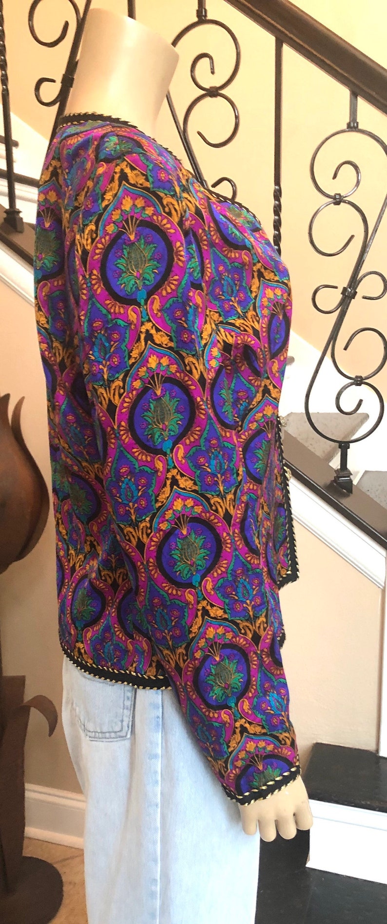 May include: A patterned jacket with a vibrant design in purple, blue, gold, and green. The jacket has a black trim and is worn over light blue jeans. The jacket's pattern features a repeating floral motif.