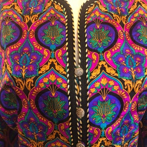 May include: A colorful patterned jacket with a black trim and silver buttons. The jacket features a vibrant design with purple, blue, green, and gold floral motifs. The trim is black with gold stitching, and the jacket has a V-neck.