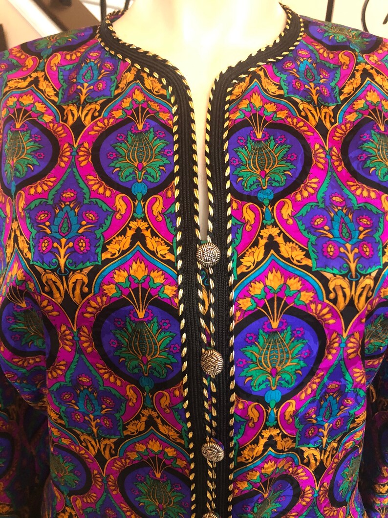 May include: A colorful patterned jacket with a black trim and silver buttons. The jacket features a vibrant design with purple, blue, green, and gold floral motifs. The trim is black with gold stitching, and the jacket has a V-neck.
