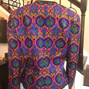 May include: A vintage jacket with a vibrant, ornate pattern in shades of purple, blue, green, and gold on a black background. The jacket has long sleeves and a black trim along the collar and hem. The back of the jacket is visible.