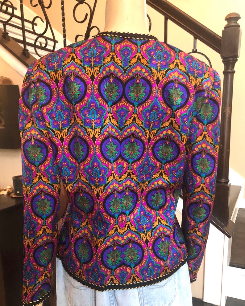 May include: A vintage jacket with a vibrant, ornate pattern in shades of purple, blue, green, and gold on a black background. The jacket has long sleeves and a black trim along the collar and hem. The back of the jacket is visible.
