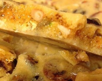 May include: Close-up of a broken piece of brittle candy with a golden brown color and a variety of nuts and seeds visible.