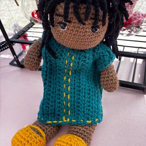 May include: A handmade crochet doll with brown skin, black yarn hair, and a small purple flower accessory. The doll wears a teal dress featuring vertical yellow stitching details and bright yellow crochet shoes. The doll has dark eyes and eyebrows.