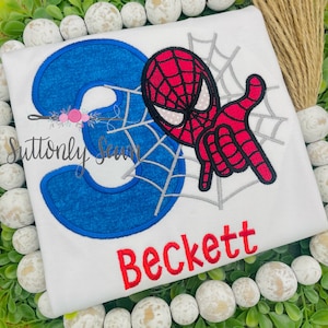 Puede incluir: Una camiseta blanca con un número 3 azul, un diseño de Spiderman rojo y negro, y el nombre Beckett en letras rojas.