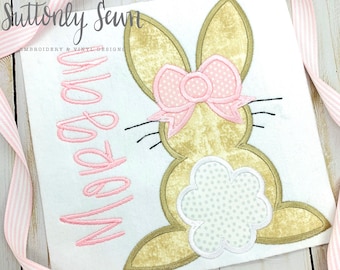 May include: A pink and gold embroidered bunny with a pink bow and white polka dots. The bunny is on a white fabric with the text "Made by" in pink.