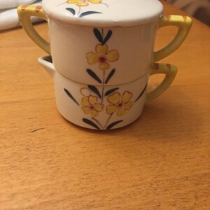 May include: A white ceramic double creamer with yellow floral designs and a lid. The creamer has two handles and is decorated with yellow flowers and green leaves.