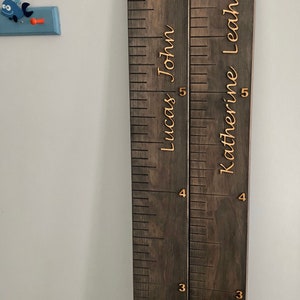 Pu&ograve; includere: Un grafico di crescita in legno con due sezioni, una per "Lucas John" e una per "Katherine Leah". Il grafico &egrave; dipinto di marrone scuro e ha numeri e nomi dorati.
