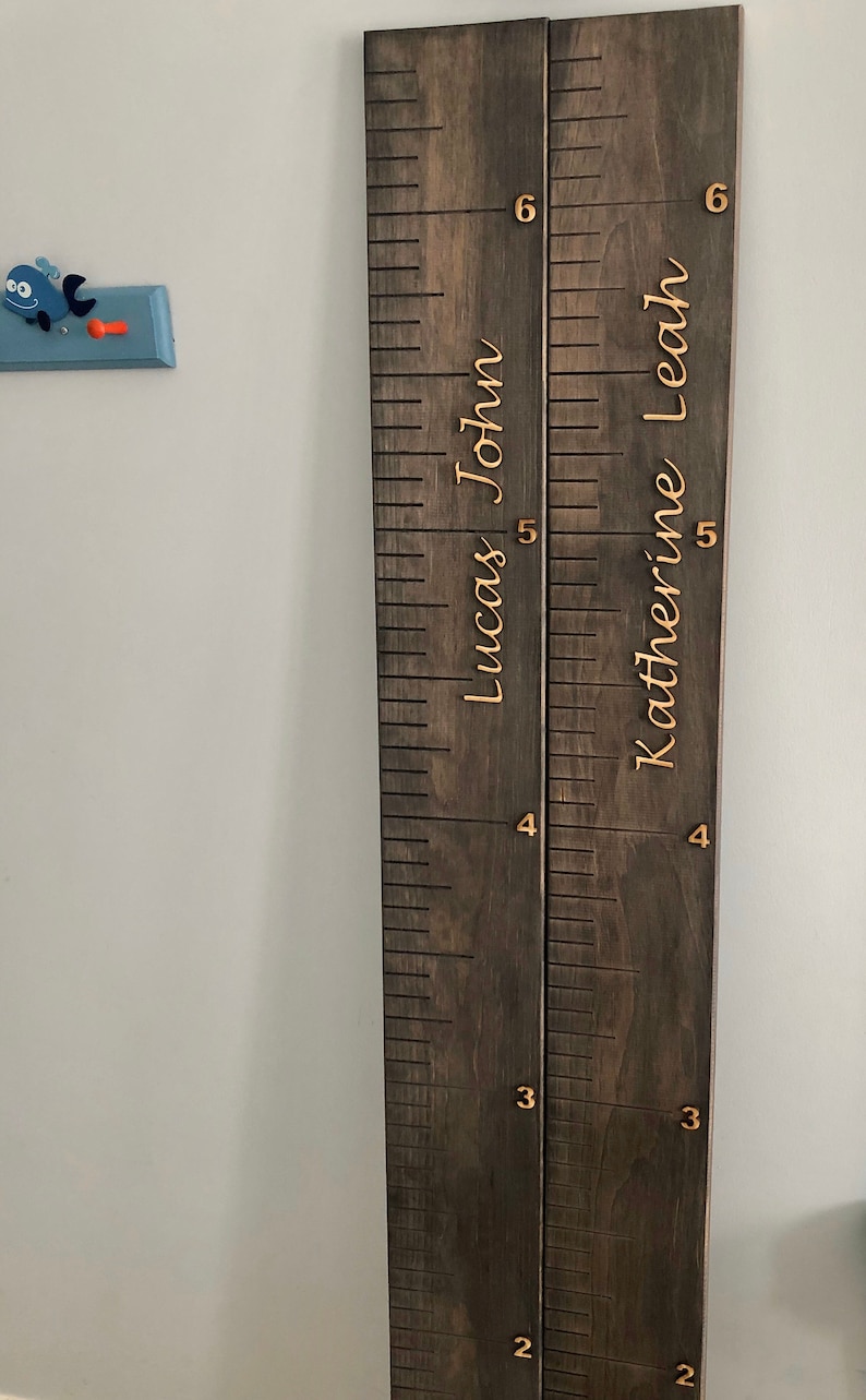 Pu&ograve; includere: Un grafico di crescita in legno con due sezioni, una per "Lucas John" e una per "Katherine Leah". Il grafico &egrave; dipinto di marrone scuro e ha numeri e nomi dorati.