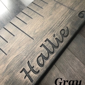 Pu&ograve; includere: Un grafico di crescita in legno grigio con segni neri e il nome "Hallie" inciso in un carattere corsivo.