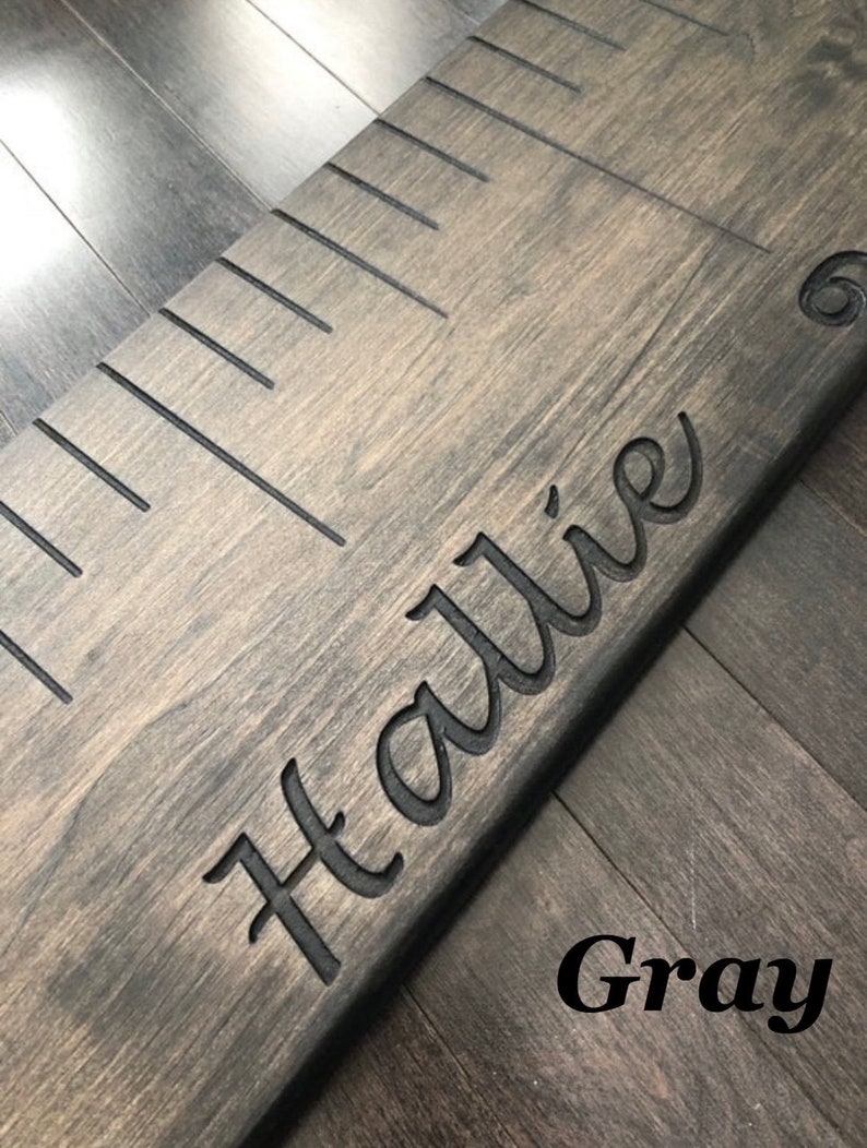Pu&ograve; includere: Un grafico di crescita in legno grigio con segni neri e il nome "Hallie" inciso in un carattere corsivo.