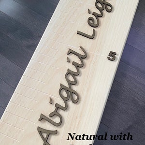 Pu&ograve; includere: Un grafico di crescita in legno naturale con un nome personalizzato "Abigail Leigh" in lettere corsive marroni scure. Il grafico ha segni per le misure di altezza.