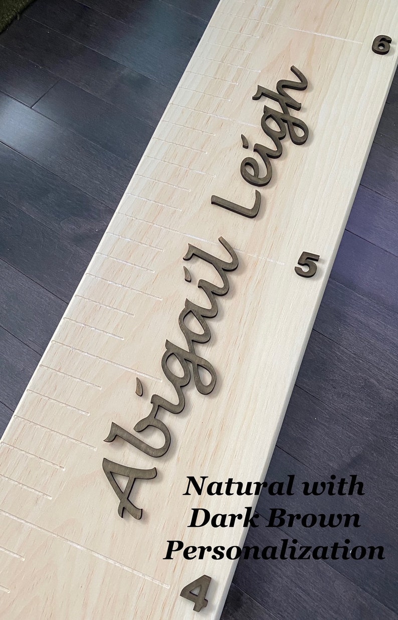 Pu&ograve; includere: Un grafico di crescita in legno naturale con un nome personalizzato "Abigail Leigh" in lettere corsive marroni scure. Il grafico ha segni per le misure di altezza.