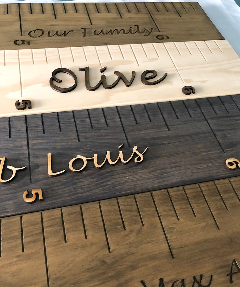 Pu&ograve; includere: Un grafico di crescita in legno con le parole "Our Family" in alto. Il grafico &egrave; composto da tre sezioni, ciascuna con una diversa tinta di legno. La sezione superiore &egrave; in legno chiaro con il nome "Olive" in corsivo. La sezione centrale &egrave; in legno scuro con il nome "Louis" in corsivo. La sezione inferiore &egrave; in legno marrone medio con il nome "Max" in corsivo.