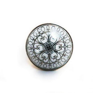 May include: A white ceramic knob with a black and silver decorative design. The knob is round and has a smooth surface.