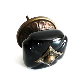 Może przedstawiać: Czarny ceramiczny gałka do szafki z mosiężną podstawą i ozdobnym mosiężnym akcentem. Gałka ma kwadratowy kształt ze zaokrąglonymi krawędziami.