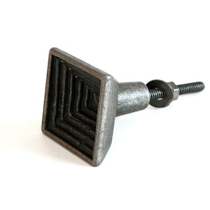 May include: A square-shaped cabinet knob with a textured design, made of metal in a dark grey finish. The knob has a screw and a washer for installation.