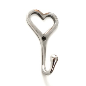 May include: A silver metal wall hook shaped like a heart. The hook has a curved end and a small hole for mounting. The heart shape is open in the middle. Ideal for hanging items.