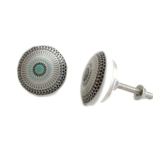 Op de afbeelding: Een witte keramische kastknop met een geometrisch ontwerp in zwart en grijs en een turquoise blauwe cirkel in het midden. De knop is bevestigd aan een zilveren schroef.
