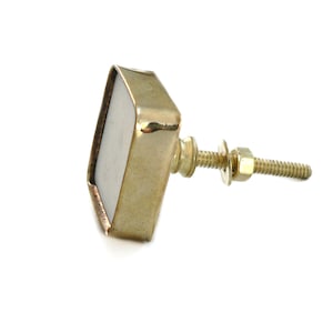 Könnte beinhalten: Goldfarbener quadratischer Schrankknopf mit weißem Zentrum und einer Schraube und Unterlegscheibe.