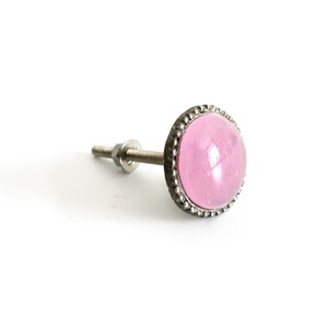Könnte beinhalten: Ein rosa Glasknopf mit einem silbernen Metallsockel und einem dekorativen Rand. Der Knopf ist an einer Schraube befestigt.