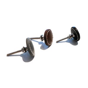 Może przedstawiać: Trzy skórzane uchwyty do szuflad ze skórzanym szwem. Uchwyty są w kolorze brązowym, szarym i czarnym.