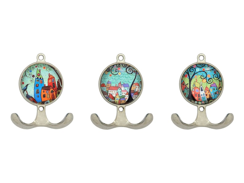 以下が含まれることがあります： 3つの装飾的な金属製の壁掛けフックのセット。各フックは、気まぐれな街のシーンの丸くカラフルなイラストが特徴です。フックはシルバー仕上げで、コートやアクセサリーを掛けるのに最適なダブルフックデザインです。
