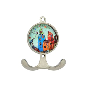 以下が含まれることがあります： 丸くカラフルなイラストが描かれた、シルバーカラーのダブルフックの壁掛け。イラストには、青、オレンジ、赤の気まぐれな家々が描かれており、青い鳥が枝にとまっています。背景は、渦巻く緑色のディテールが施された水色です。