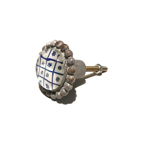 Könnte beinhalten: Weißer Keramik-Knopf mit blau-weißem Muster. Der Knopf hat eine silberne Metallbasis und einen perlenbesetzten Rand.