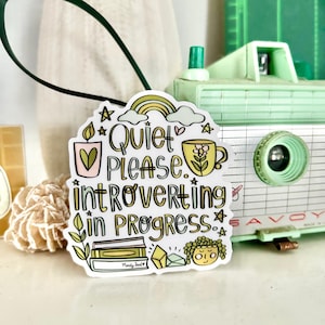 May include: White sticker with a colorful illustration of a cup of tea, a rainbow, stars, and the text "Quiet Please. Introverting in Progress."