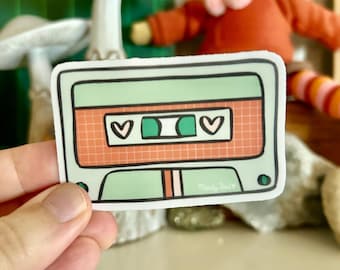 May include: A green and orange cassette tape sticker with a grid pattern and two hearts on the tape. The sticker has the text "Mandy Joel" in the bottom right corner.