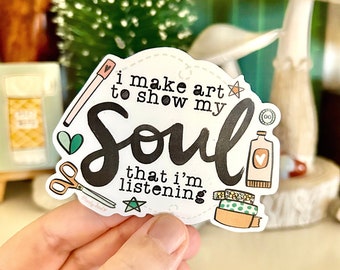 May include: White sticker with black text that reads "I make art to show my soul that I'm listening". The sticker has various illustrations including a heart, scissors, a star, and a bottle of paint.