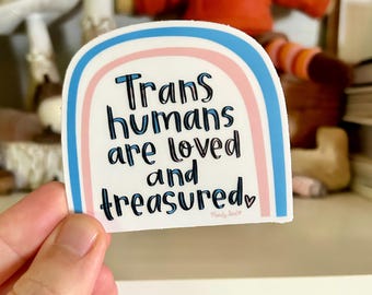 May include: A sticker with a rainbow design in blue, pink, and white, with the words "Trans humans are loved and treasured" in black lettering. The sticker has a white background and a rounded top.