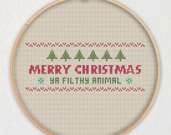 May include: A cross-stitch pattern on a beige fabric stretched over a wooden embroidery hoop. The pattern features a red and white border, six green Christmas trees, and the text "Merry Christmas Ya Filthy Animal".
