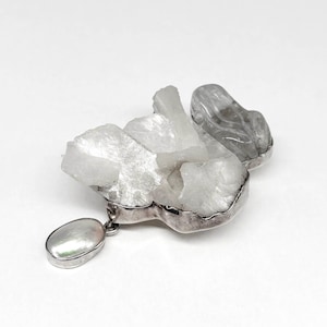May include: A silver-toned pendant featuring a cluster of white, crystalline formations. A pearlescent, oval-shaped charm dangles from the bottom. The pendant has a silver border and is set against a white background.