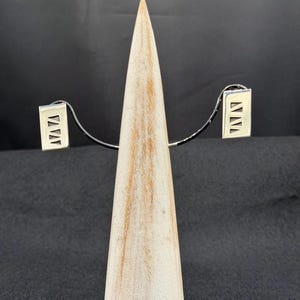 May include: A pair of rectangular silver earrings featuring a cut-out design of three stacked triangles. The earrings are displayed on a light-colored wooden cone stand, showcasing a polished metallic finish and a geometric pattern on each piece.