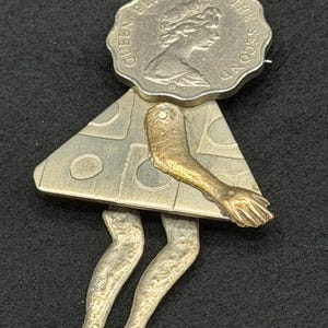 May include: A brooch shaped like a figure, featuring a scalloped silver coin head embossed with Queen Elizabeth the Second. The body is a triangular silver piece with circular indentations, a single brass-toned arm, and two silver legs.