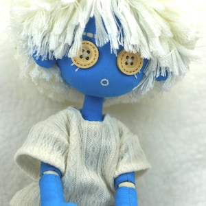 Op de afbeelding: Handgemaakte pop met een blauw gezicht en ledematen, knoopogen en wit garen haar. De pop draagt een witte trui en lichtblauwe shorts. De pop zit.