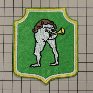 Op de afbeelding: Geborduurde patch met een grillige figuur die op een gouden hoorn blaast. De figuur is wit met bruin haar, tegen een groene schildvormige achtergrond met een gele rand. Een uniek, decoratief item.