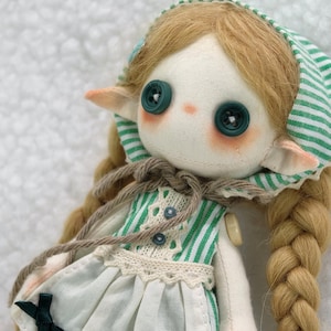 Puede incluir: Muñeca hecha a mano con ojos de botón y pelo largo trenzado. La muñeca lleva un vestido a rayas verdes y blancas con un delantal blanco y lazos verde oscuro. La cara de la muñeca es pálida con mejillas rosadas y orejas pequeñas y puntiagudas.