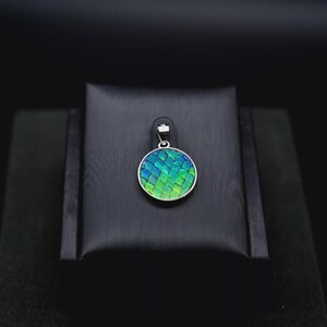 May include: A circular pendant with a silver-coloured frame. The pendant has a scale-like pattern in shades of blue and green. The pendant is displayed on a black stand against a black background.