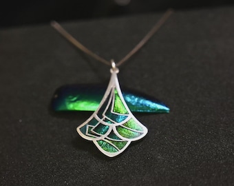 Op de afbeelding: Een zilveren hangerketting met een geometrisch ontwerp. De hanger heeft een waaiervormig ontwerp met groene en blauwe emaille details. De ketting wordt gepresenteerd op een zwart oppervlak.